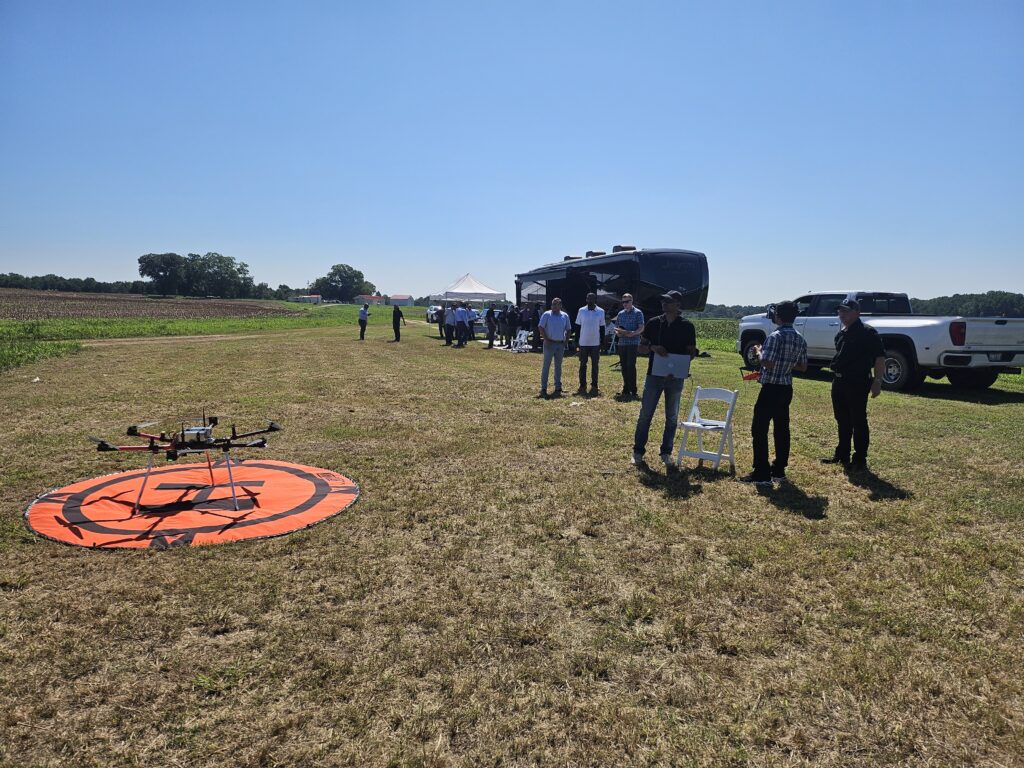 A pilot prepares to fly a drone in a fields with several other people and vehicles in the vicinity.