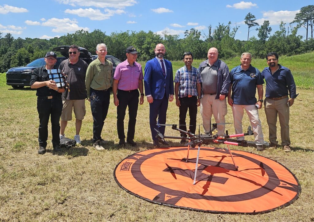 A group of men stand for a photo behind a drone on a landing pad in a field.